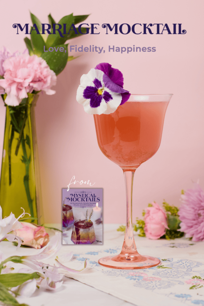 Pink stemmed cocktail glass filled with a nonalcoholic wedding mocktail (called Marriage Mocktail) garnished with a purple and white edible flower.