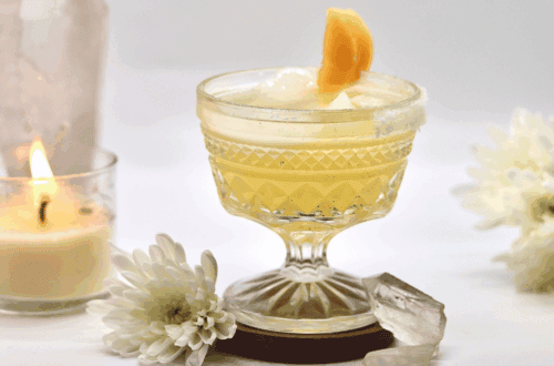 Melon-Yellow Imbolc drink recipe in short stemmed cocktail glass with moon-shaped melon garnish. Clear quartz crystals, white flower, and candle in the background.