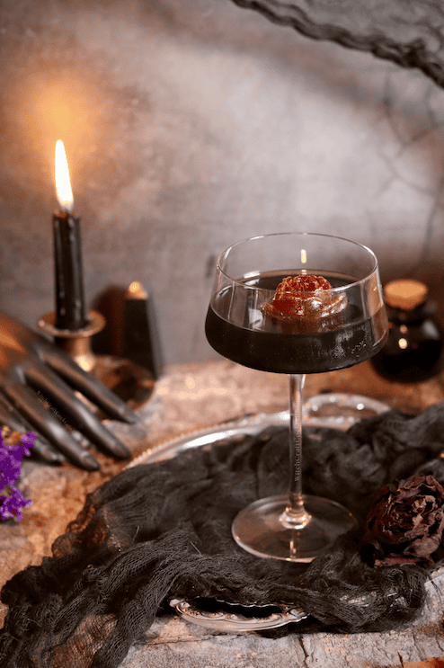 Closeup of the Black Heart drink, showcasing the black color of the cocktail accented by a striking red rose ice cube garnish against a grey backdrop and candle.