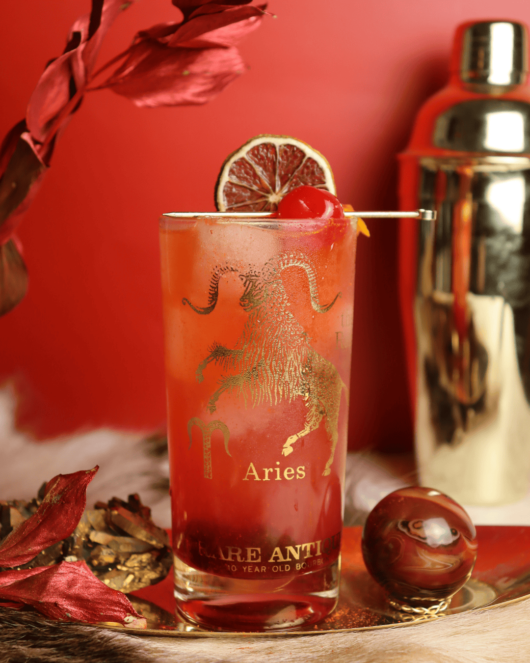 Red Aries cocktail in collins glass against a red background with gold accents.