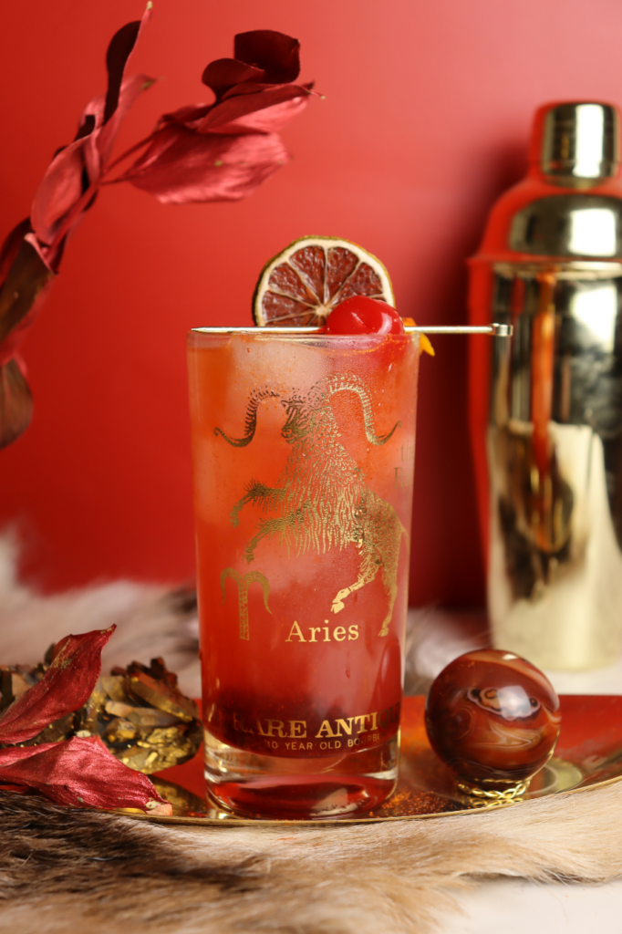 Red Aries cocktail in collins glass against a red background with gold accents.