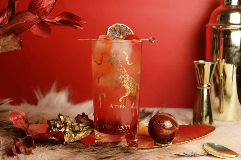 Red Aries cocktail in collins glass against a red background with gold accents.