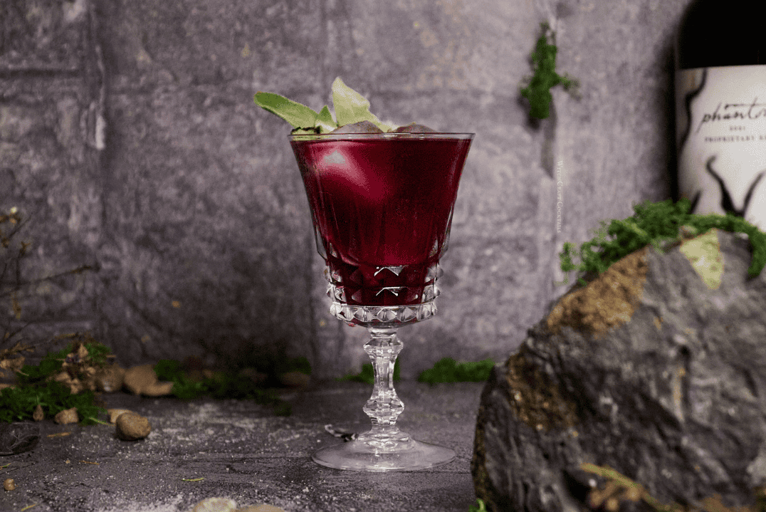 Red sangria with sage garnish in fancy glassware.